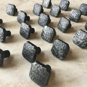 May include: A collection of 20 square-shaped cabinet knobs with a hammered metal finish. The knobs are black with a silver-grey patina.