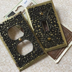 May include: Two vintage electrical outlet and switch plates with ornate black filigree designs on a gold background. The plates are new, still in their original packaging from Sears.