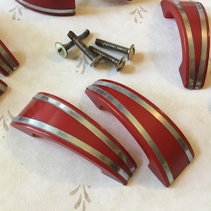 May include: Red and silver cabinet pulls with a curved design. The pulls have a glossy finish and are accented with two silver stripes. Several pulls and screws are scattered on a patterned surface.