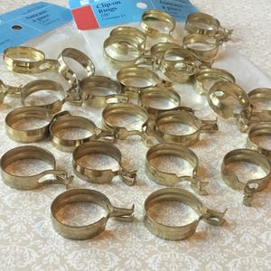 Peut inclure: Plusieurs anneaux à clipser de couleur laiton, chacun avec un levier pour l'ouverture et la fermeture. Les anneaux sont dispersés sur une surface à motifs, avec deux paquets d'anneaux à clipser en arrière-plan. Les paquets indiquent "Clip-on Rings 7/8" et "Contains 14".