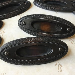 May include: A collection of antique black oval drawer pulls with floral and beaded detailing. Each pull features a recessed centre and is fixed with two screws. The metal pulls have a distressed finish, typical of vintage hardware.