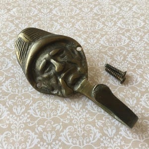 May include: Antique brass wall hook with a detailed face design. The hook has a curved shape and includes two small screws for mounting. The brass has a weathered appearance, adding to its vintage charm. A decorative item for hanging items.