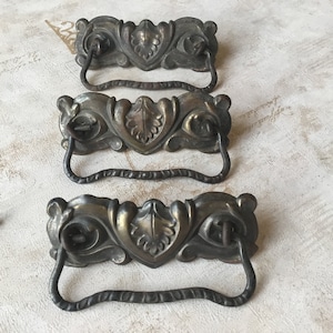 Op de afbeelding: Drie antieke messing ladegrepen met sierlijke bladmotieven en een gebogen handvat. De grepen zijn in een rij op een wit oppervlak gerangschikt.