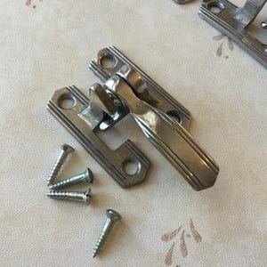 May include: A silver-toned metal latch with a lever handle and decorative grooves. The latch is attached to a surface with screws.