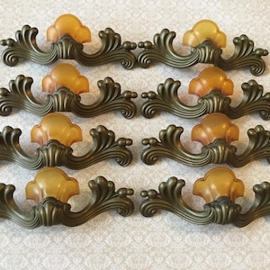 May include: Eight antique bronze-toned drawer pulls with amber-colored center accents. The pulls have a decorative, ornate design with curved lines and floral-like details, perfect for vintage furniture restoration.