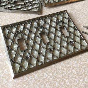 Könnte beinhalten: Ein Satz Vintage-Lichtschalterabdeckungen aus silberfarbenem Metall. Die Abdeckungen haben ein Rautenmuster und rechteckige Schalteröffnungen. Mehrere Schrauben sind in der Nähe verstreut.