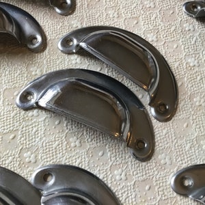 May include: A set of six silver-toned metal cabinet drawer pulls. The pulls are shaped like a half-circle with a slight curve in the centre. They have a smooth, polished finish.