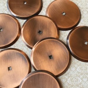 May include: A collection of eight round, copper-colored metal cabinet hardware pieces. Each piece has a square hole in the center. The hardware has a brushed finish, and the color varies slightly across the pieces.