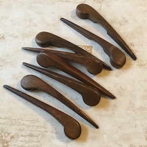 May include: A set of eight dark brown wooden hair clips. The clips are long and slender with a rounded end. They are arranged in a fan shape on a light brown surface.