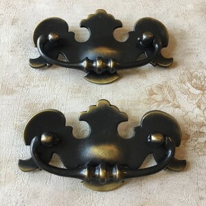 May include: A pair of antique-style drawer pulls in dark bronze with brass highlights. Each pull has a decorative backplate, a curved handle, and two small knobs. Ideal for furniture restoration or DIY projects, these pulls are a classic design.