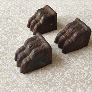 Op de afbeelding: Drie antieke meubelpoten van gietijzer, elk in de vorm van een klauw. De poten zijn donkerbruin van kleur met een getextureerd, verouderd uiterlijk. Ze zijn gerangschikt op een crèmekleurig oppervlak met een bloemmotief.