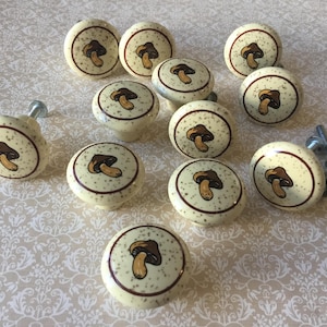 May include: A collection of cream-colored mushroom-themed cabinet knobs. Each knob features a brown mushroom illustration in the center, surrounded by a brown border and speckled design. Several silver screws are also visible.