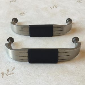 Puede incluir: Dos tiradores de puerta de armario plateados con insertos rectangulares negros. Los tiradores son curvos y tienen un diseño moderno.