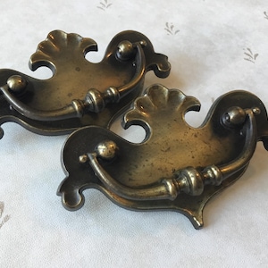 Peut inclure: Deux poignées de tiroir en laiton antique avec des motifs ornés et enroulés. Les poignées ont une poignée en boucle et sont en finition vieillie.