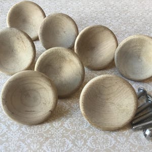 May include: A collection of eight unfinished wooden cabinet knobs with a rounded design. The knobs are light beige in color and have visible wood grain. Several silver screws are also included.