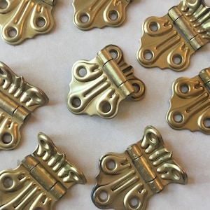 Puede incluir: Ocho bisagras de metal dorado antiguo con diseños ornamentados. Las bisagras tienen forma de mariposa con un centro decorativo y dos pequeños bucles a cada lado.