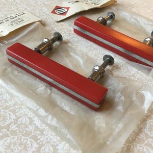 May include: Three vintage red and silver cabinet pulls with silver hardware, still in their original packaging. The pulls are rectangular with a silver stripe down the center. The packaging has the text "Building Material Center" and "Harvest of Polo Park".