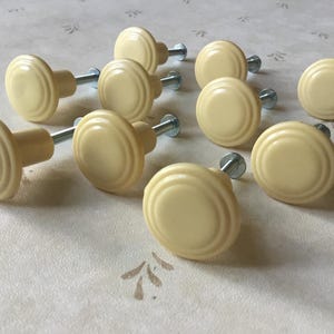 May include: A set of ten cream-coloured cabinet knobs with a ridged design. Each knob has a round face and a metal screw for attachment. The knobs are arranged on a patterned surface.