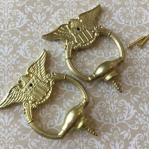 Pode incluir: Duas puxadores de gaveta dourados em estilo vintage com um design de águia e escudo. Cada puxador tem uma alça de anel e um parafuso para montagem. Dois parafusos adicionais também são visíveis.