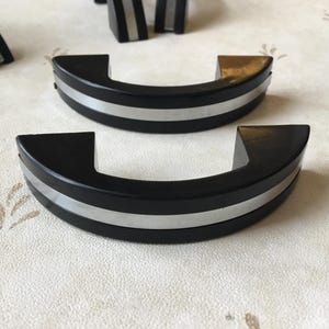 Puede incluir: Dos tiradores de puerta de armario negros con una franja plateada. Los tiradores son curvos y tienen un diseño moderno.