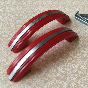 Peut inclure: Deux poignées de meuble rouges avec une bande argentée et trois vis argentées. Les poignées ont un design incurvé et sont faites d'un matériau brillant. La quincaillerie est posée sur un fond à motifs.