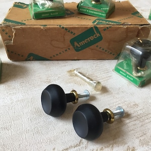 Pode incluir: Um close-up de duas maçanetas pretas para armários com detalhes dourados. As maçanetas estão embaladas em plástico transparente e etiquetadas com "Amerock Genuine Hardware".