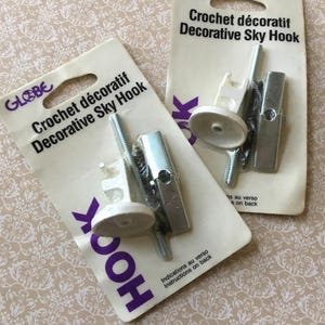 May include: Two packs of "Crochet décoratif Decorative Sky Hook" hardware. Each pack contains a white hook, a metal bracket, and screws. The packaging is white with purple text and a floral background.