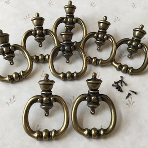 May include: Eight antique brass cabinet knobs with a decorative design. The knobs have a ring pull and a small finial on top. The knobs are accompanied by eight small screws.