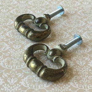 以下が含まれることがあります： アンティークスタイルの引き出し用つまみ2つ。使い古したゴールド仕上げで、それぞれ曲線的なハート型デザイン、装飾的なベース、取り付け用のシルバースクリューが付いています。ハードウェアは模様入りの背景に配置されています。