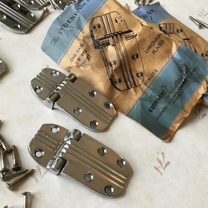 May include: Two pairs of chrome plated hinges with a decorative design. The hinges are packaged in a blue and white box with the text "The Sydenham" and "Cuthbert Industries Ltd."