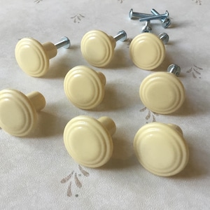 May include: Nine cream-coloured cabinet knobs with a ridged design. Each knob has a metal screw attached. Several silver screws are also visible, suggesting a hardware set for furniture restoration or new construction.