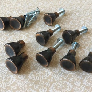May include: A collection of bronze-colored cabinet knobs with silver screws. The knobs have a rounded, bell-shaped design with a dark finish and a metallic sheen. The hardware is arranged on a patterned surface.