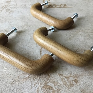 May include: Three wooden cabinet knobs with a curved design and silver screws. The knobs are made of light brown wood with a natural grain pattern.