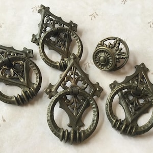 May include: Five antique cabinet knobs with ornate designs. The knobs are made of metal and have a dark brown finish. Four of the knobs are ring pulls with a square frame and a circular pull. The fifth knob is a round knob with a floral design.