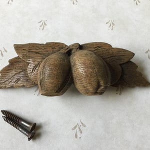 Pode incluir: Um elemento decorativo em madeira entalhada com duas formas em forma de bolota com detalhes de folhas. A peça é feita de madeira castanha clara e inclui dois parafusos.