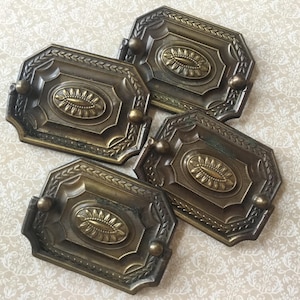 May include: Four antique bronze drawer pulls with an oval center design and decorative leaf accents. Each pull has a small knob on each end. The pulls are rectangular with an octagonal shape.