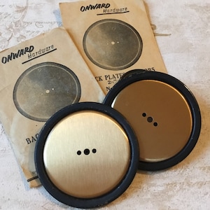 Op de afbeelding: Twee goudkleurige metalen achterplaten met zwarte rubberen ringen. De platen hebben elk drie gaten en worden geleverd met instructies van Onward Hardware.