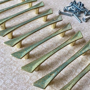 May include: A set of eight cabinet pulls with a distressed teal and gold finish. The pulls have a unique, angular design and are accompanied by a pile of silver screws. The hardware is likely for furniture restoration or new construction.