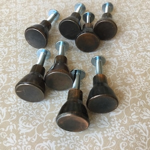 Peut inclure: Huit boutons de meuble de couleur bronze avec des vis argentées. Les boutons ont un dessus rond et plat et une base conique. Ils sont disposés sur une surface beige à motifs.