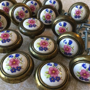 May include: A collection of antique brass cabinet knobs with white ceramic faces. Each knob features a floral design with pink, blue, and green accents. The knobs are round and have a vintage aesthetic, with screws included.