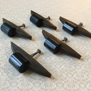 Może przedstawiać: Pięć uchwytów do szafek w stylu vintage w kolorze czarnym i brązowym. Każdy uchwyt ma cylindryczny czarny uchwyt i brązową podstawę w kształcie skrzydła, przymocowaną srebrną śrubą. Uchwyty są ułożone na wzorzystej, kremowej powierzchni.