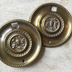 Op de afbeelding: Twee antieke messing deurknopjes met een eikel- en eikenbladontwerp. De knoppen hebben een touwachtige rand en een sleutelgat.