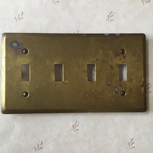 Peut inclure: Plaque d'interrupteur en laiton antique avec quatre ouvertures pour interrupteurs. La plaque rectangulaire présente une finition légèrement ternie et est fixée avec quatre vis. Le fond est un papier peint à motifs avec de petits motifs en forme de feuilles.