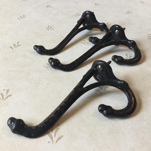 May include: Three antique black metal coat hooks. Each hook has a curved design with a rounded end and a decorative top. The hooks are arranged on a light-colored surface with a subtle pattern.