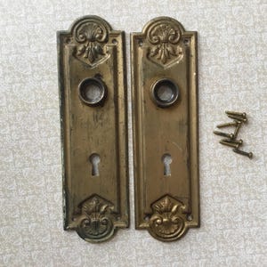 Peut inclure: Deux plaques de porte anciennes en laiton avec trous de serrure et ornements floraux décoratifs. Les plaques sont rectangulaires avec une ouverture circulaire pour la poignée et un trou de serrure en dessous. Plusieurs petites vis en laiton sont incluses.