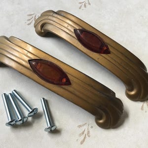 Op de afbeelding: Twee antieke goudkleurige kastgrepen met een decoratief ontwerp en een rood-oranje accent. Inclusief zilveren schroeven voor installatie. Deze vintage handgrepen zijn een decoratief hardware-element.