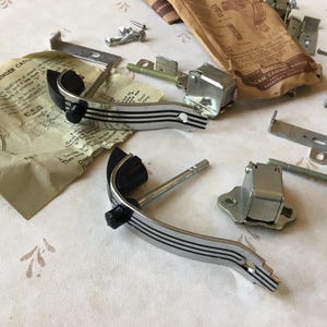 May include: Two silver and black cabinet door catches with instructions on how to install plunger catches. The catches have a silver finish with black plastic knobs. The instructions are printed on a white paper with black text.