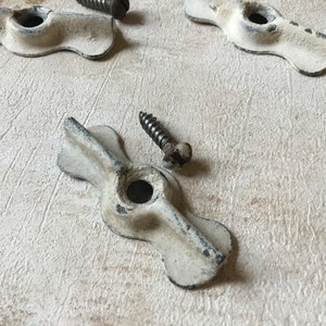 May include: Close-up of several antique white metal drawer pulls with a distressed finish, each with a central hole and two screw holes.  Dark screws are scattered around the pulls, suggesting a vintage hardware set.