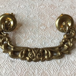 May include: An ornate metal drawer pull in a gold tone, featuring a decorative scroll design. The pull has two round mounting plates with screw holes.
