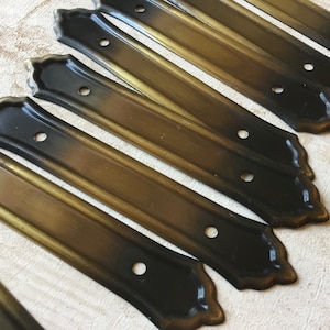 May include: A set of eight antique-style metal drawer pulls with a black and gold finish. The pulls have a decorative, scalloped edge and two holes for mounting.